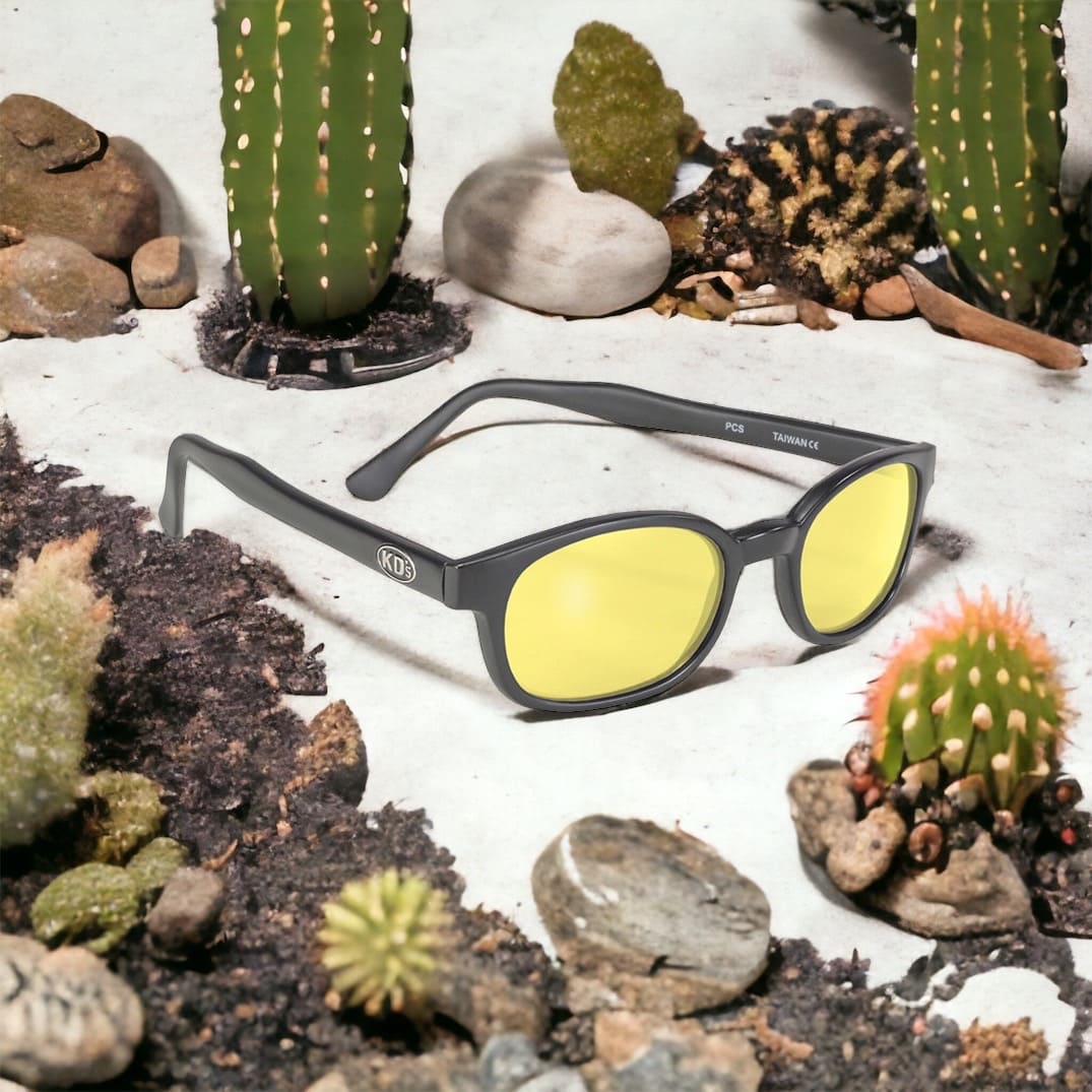 KD's 21112 sunglasses that fit under a motorcycle or ski helmet. For outdoor activities. With a durable matte black frame and yellow tinted lenses. Sitting in the sand surrounded by cacti.