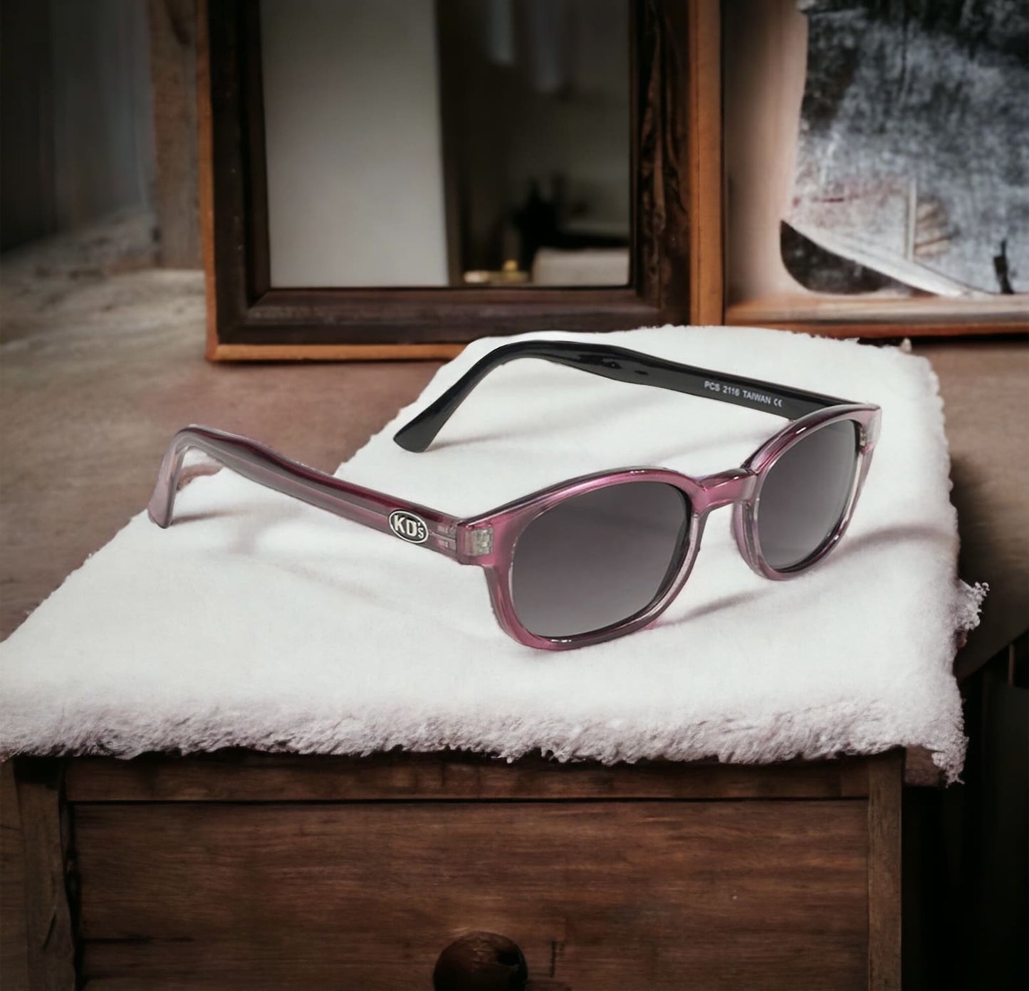 The KD's 2116 sunglasses perfect for sunny days with grey lenses and an elegant pink frame laid on a soft blanket in a wooden house.