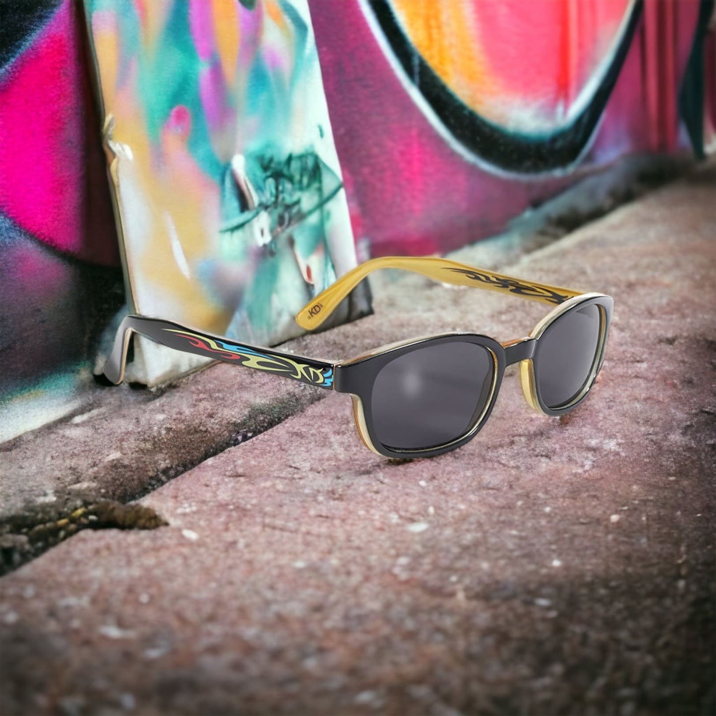 The KD's 5400 sunglasses with grey polycarbonate lenses and a black frame with a light blue and red tribal tattoo pattern exposed on a sidewalk behind graffiti.