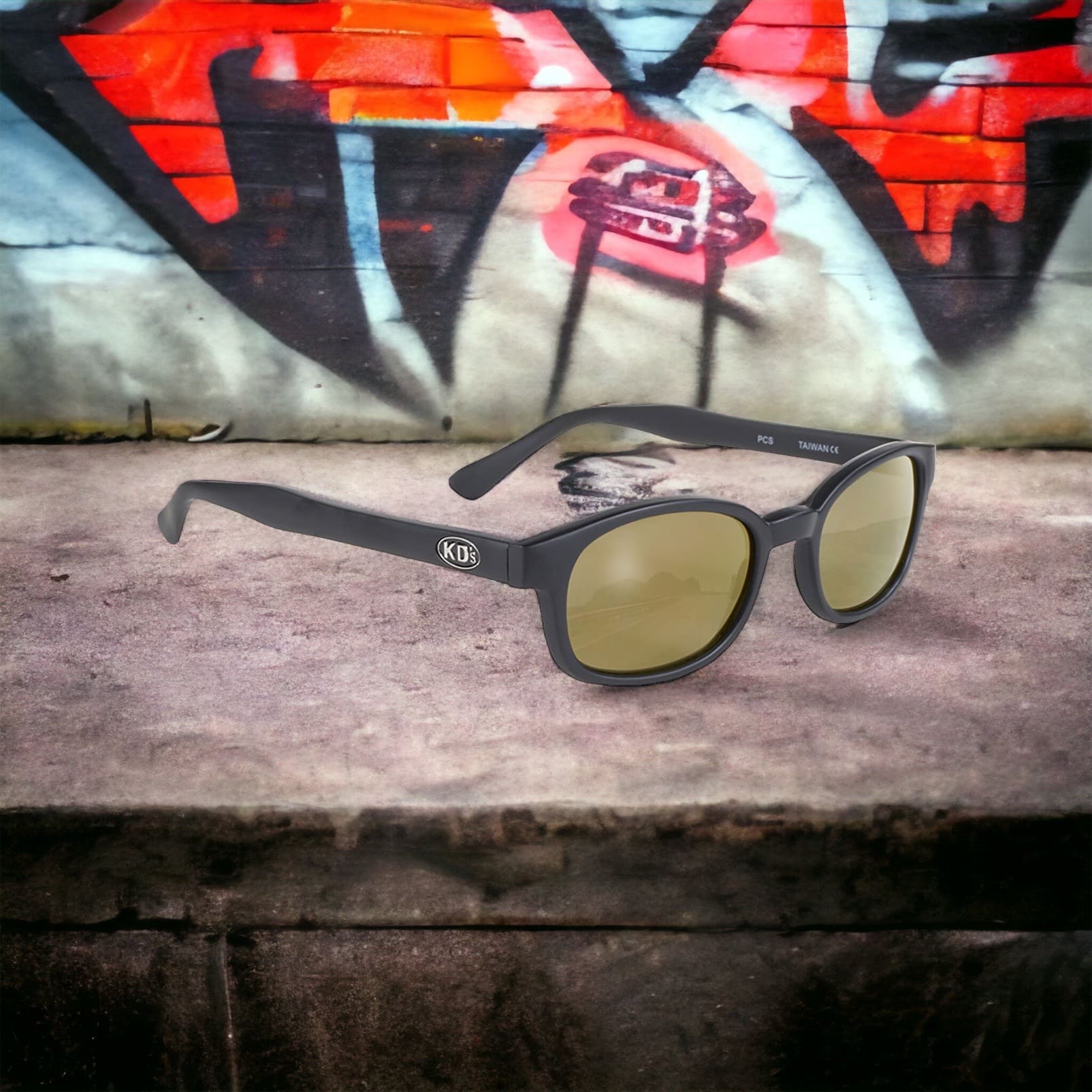 Les lunettes de soleil KD's 2000 avec des verres miroirs doré et une monture noir mat posé sur un trottoir derrière un mur rempli de graffiti.