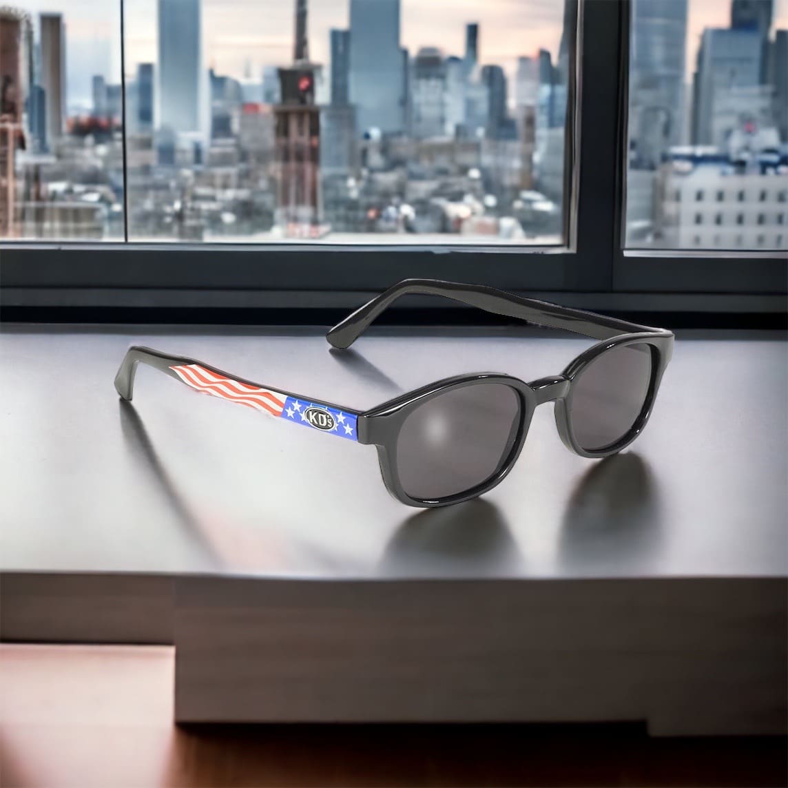 KD's 20050 sunglasses that fit under a motorcycle or ski helmet. With a sturdy frame decorated with the U.S. flag and gray tinted lenses placed in front of a window with a view of New York.