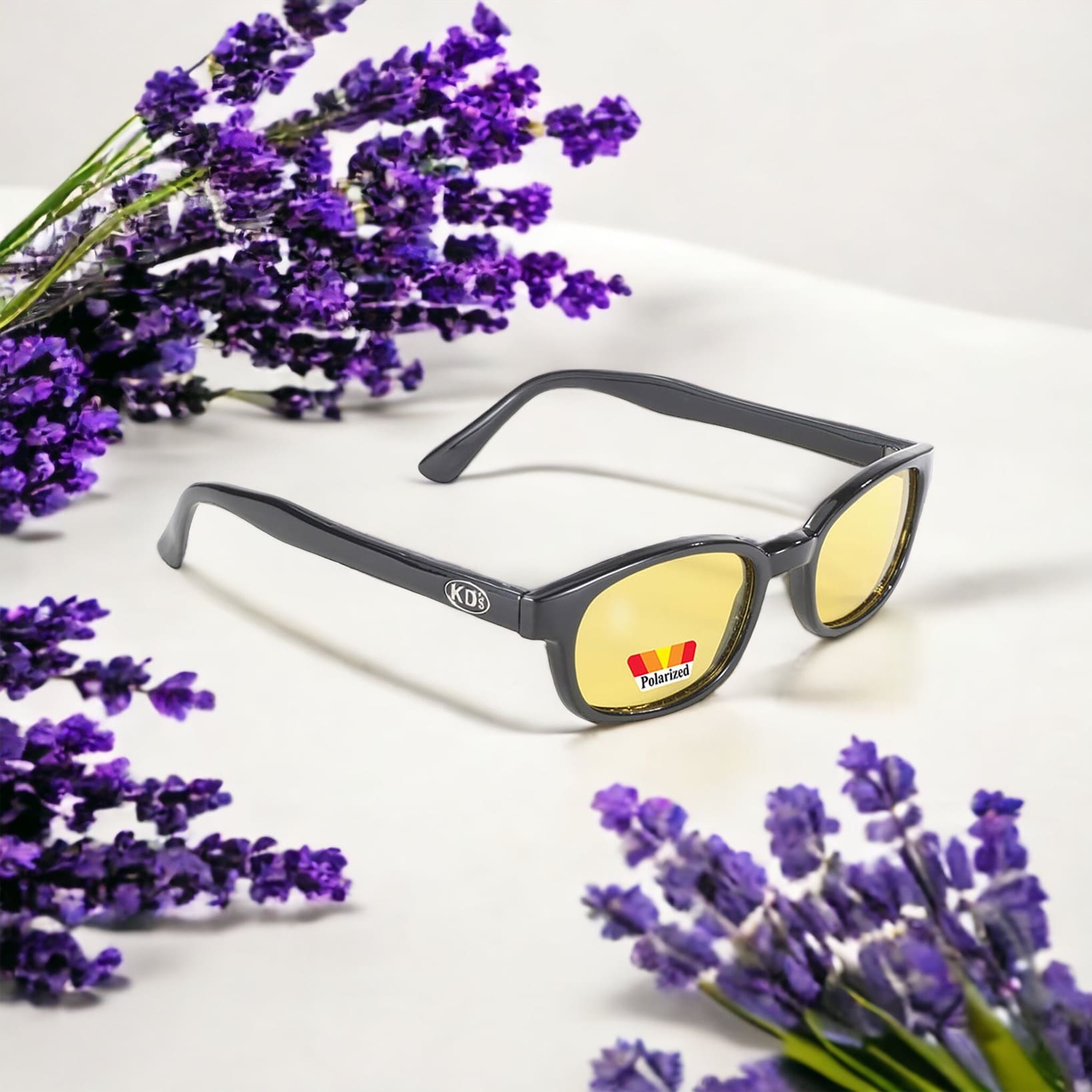 KD's 20129 sunglasses that fit under a motorcycle or ski helmet. For bikers and sportsmen. With a durable black frame and polarized yellow tinted lenses. Placed on a white table surrounded by lavender.
