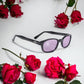 KD's 21216 sunglasses that fit under a motorcycle or ski helmet. Worn by SAMCRO bikers and Jax Teller in Sons of Anarchy. With a sleek black frame and purple tinted lenses. Set on a white surface surrounded by roses.