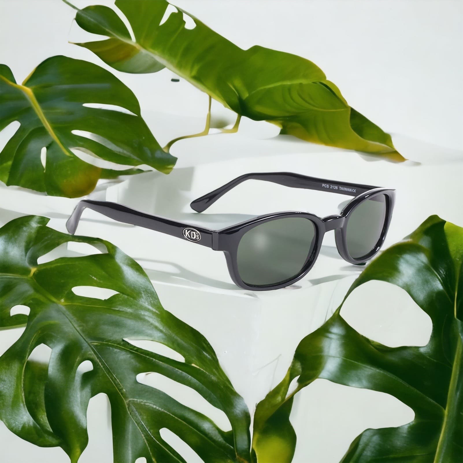 The stylish KD's 2126 sunglasses with dark green polycarbonate lenses and a solid black frame placed on a white furniture surrounded by plants.