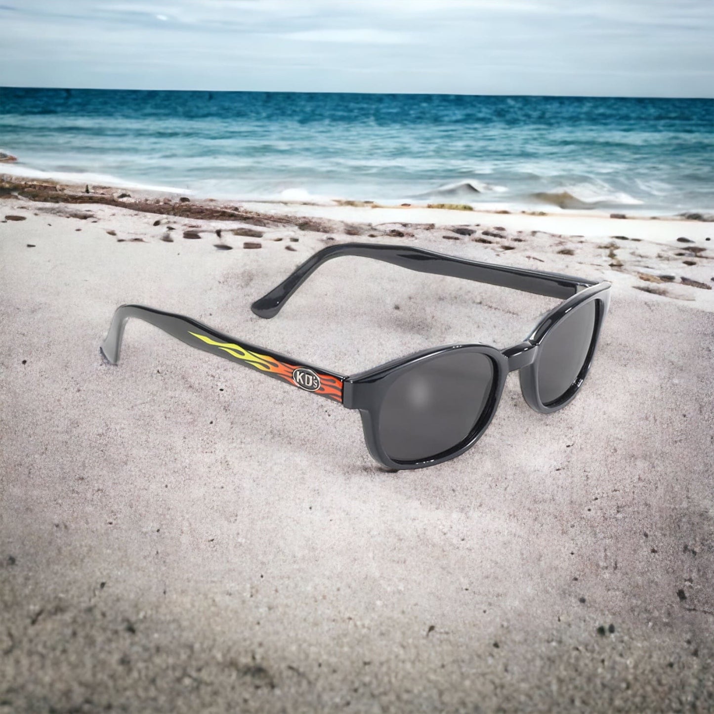 The KD's 3010 sunglasses with grey polycarbonate lenses and a frame with a yellow and orange gradient flame pattern exposed on the sand of a beach.