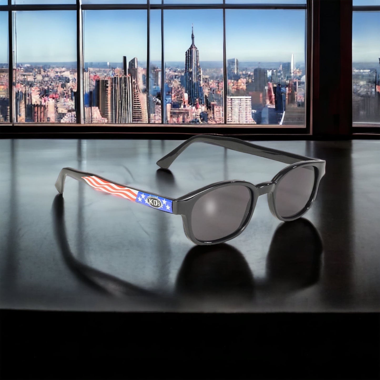 X-KD's 10050 large sunglasses with gray polycarbonate lenses and a USA flag frame set on a desk with a view of New York.