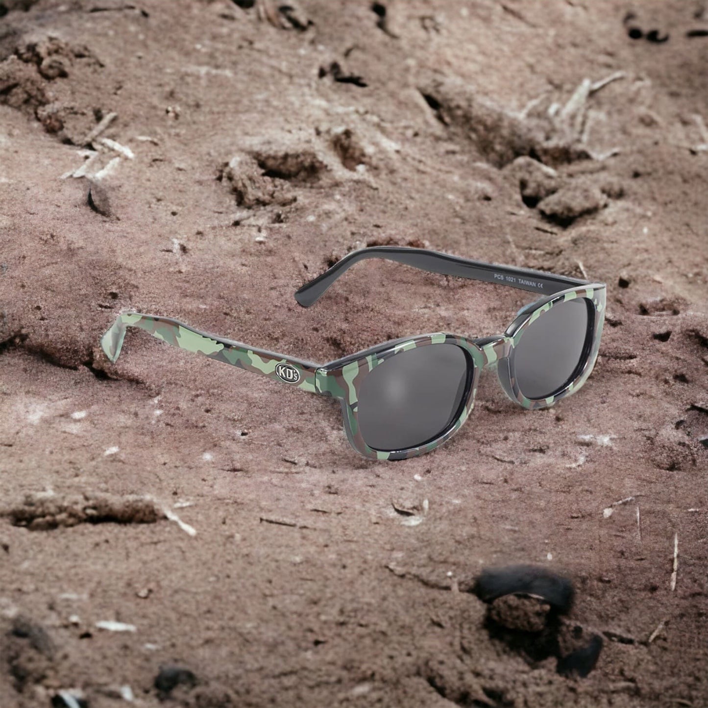 The X-KD's 1021 sunglasses with grey lenses and a military camo frame decor on the beaten earth