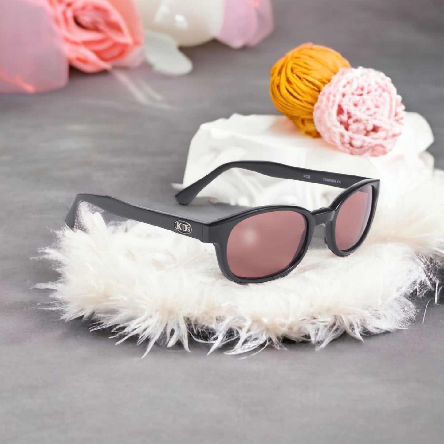 Large original X-KD's Crystal 12120 sunglasses with solid black frames and pink tinted polycarbonate lenses set on feather furniture.