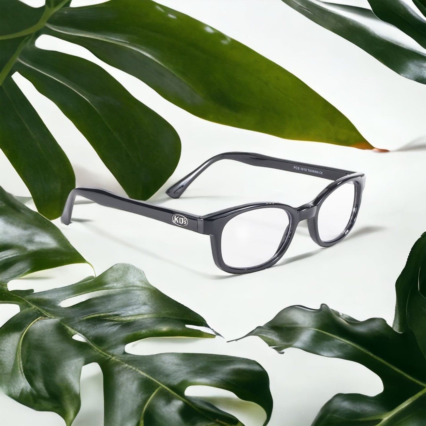 X-KD's 1015 sunglasses with transparent lenses and a sophisticated black frame placed on a white desk surrounded by leaves